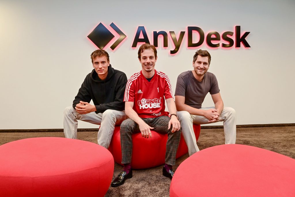 Three men are sitting in front of the AnyDesk logo. The man in the middle is wearing a red jersey.