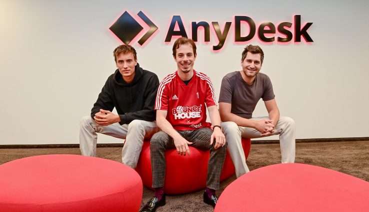 Three men are sitting in front of the AnyDesk logo. The man in the middle is wearing a red jersey.